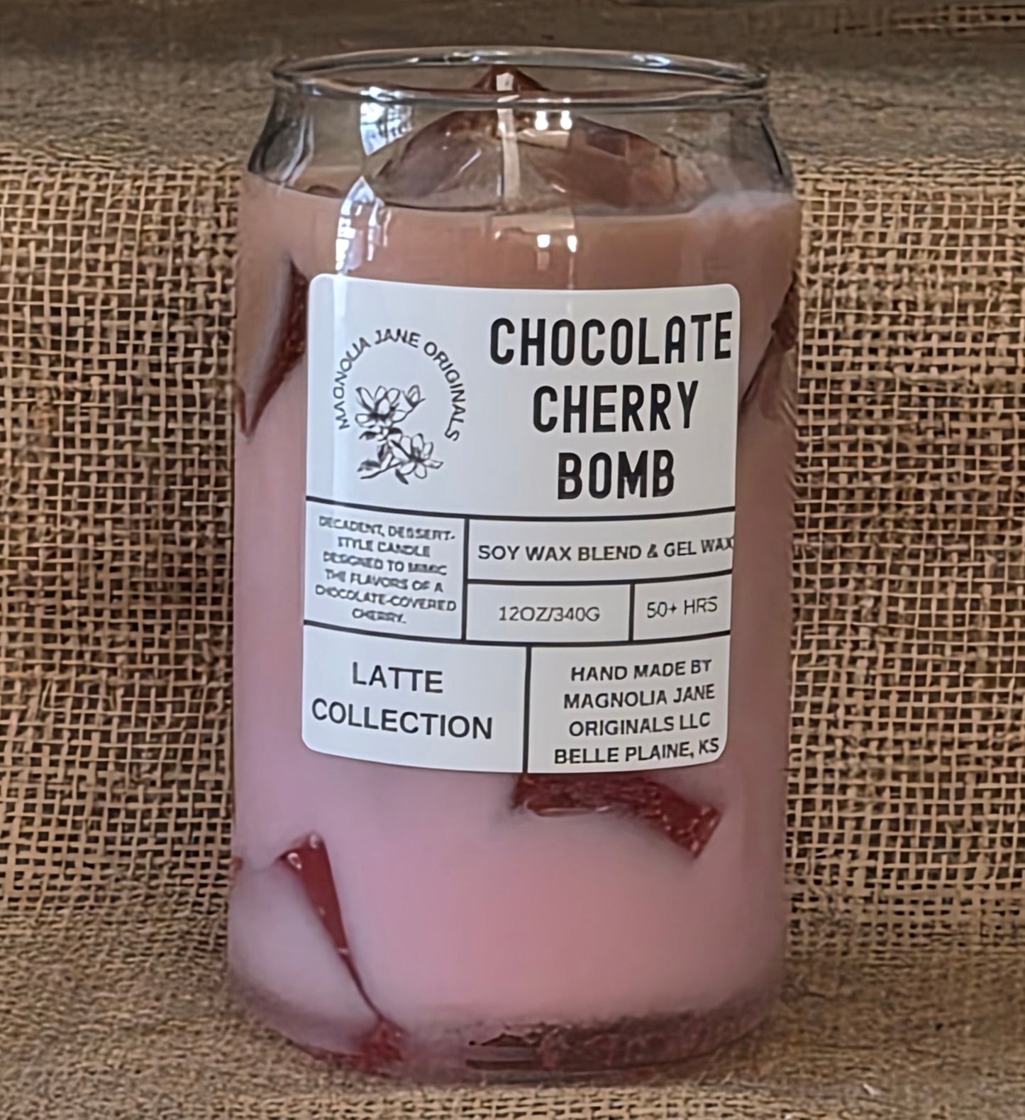 Candle in a glass jar labeled 'Chocolate Cherry Bomb' that is pink and brown with clear gel wax ice cubes from the Latte Collection on a burlap background.