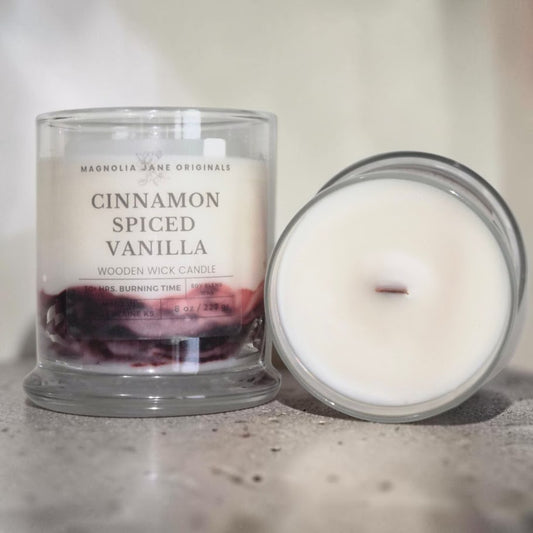 A glass jar candle labeled 'Cinnamon Spiced Vanilla' by Magnolia Jane Originals, shown next to its open lid on a neutral surface.