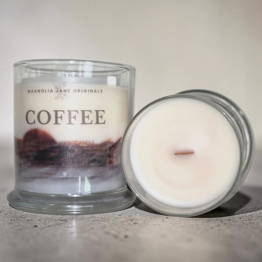 A glass jar candle labeled 'COFFEE' by Magnolia Jane Originals, shown with the lid placed beside it on a neutral surface.
