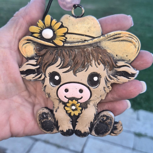 A hand holding a hand-painted cow ornament outdoors. The ornament features a light brown cow wearing a yellow hat and holding a yellow flower, with a black hanging loop at the top.