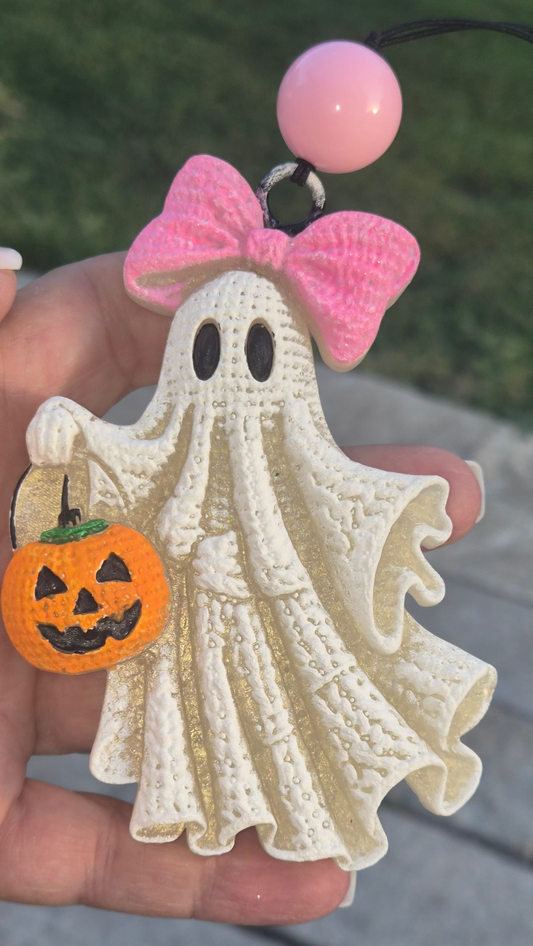 A hand holds a Halloween ghost charm with a pink bow, black eyes, and an orange jack-o'-lantern bucket. The charm has a textured cream-colored ghost body and a pink bead at the top, set against a blurred outdoor background of grass and stone pavers.
