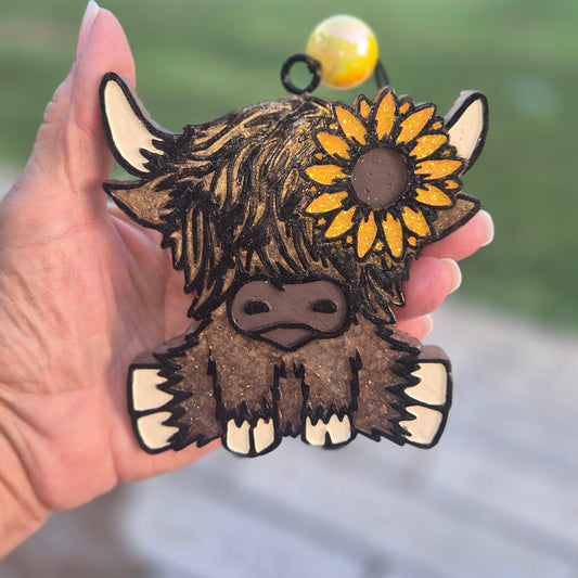 A hand holding a cartoon Highland cow ornament or charm with a yellow sunflower and a small yellow bead at the top, photographed outdoors with a blurred green lawn and stone path in the background.